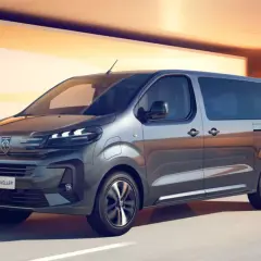 Szary bus Peugeot e-Traveller z przyciemnianymi szybami na tle pomarańczowej architektury.