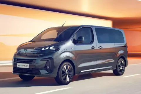 Szary bus Peugeot e-Traveller z przyciemnianymi szybami na tle pomarańczowej architektury.